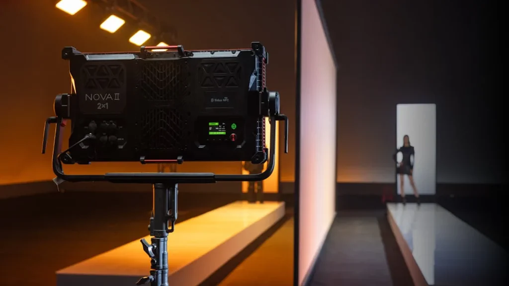 Панель Aputure Nova II 2×1 на стійці великим планом, на екрані приладу активне підключення Sidus NFC; на задньому плані тепле помаранчеве підсвічування подіуму й силует людини в чорному біля білої колони — атмосфера модного показу чи іміджевої зйомки.